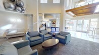 This well-appointed single-floor space combines a spacious living lounge and a fully equipped kitchen into one cohesive, open-concept layout. The living area immediately impresses with its dramatic double-height ceilings, which lend an airy, expansive feel throughout. A bold, floor-to-ceiling textured accent wall serves as the focal point of the lounge, framing a large flat-screen television and flanked by framed artwork. Multiple seating arrangements featuring contemporary gray upholstered chairs and sofas are grouped around dark wood coffee tables, creating comfortable areas for gathering or relaxation. Patterned carpet with a subtle circular motif adds visual interest underfoot. Large windows along the perimeter flood the space with natural light and offer views of the surrounding outdoors. The kitchen area is defined by a striking curved island with a dark, high-gloss finish and granite countertops, complemented by dark hardwood flooring. White cabinetry lines the walls alongside stainless steel appliances, including a refrigerator and coffee station. A large circular pendant light fixture elegantly anchors the kitchen zone. Architectural columns and curved ceiling details add a refined, modern character, while upper-level glass railings hint at the vertical scale of the space. Overall, the design balances contemporary aesthetics with a welcoming, functional atmosphere.