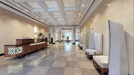 This expansive lobby presents an impressive first impression through a carefully curated blend of classic architecture and contemporary design. Upon entry, polished terrazzo floors in a checkered pattern extend throughout the space, reflecting ambient light and reinforcing the refined aesthetic. The main reception area features a handsome wood-paneled desk accented with decorative lattice detailing, set beneath dramatically high coffered ceilings fitted with recessed lighting that illuminates warm, stone-clad walls.

A central seating lounge offers a circular upholstered bench and cushioned chairs arranged around a sculptural centerpiece, with tall windows framing lush greenery beyond. Along one wall, a series of high-backed upholstered privacy chairs are positioned beside vertical ribbed decorative panels, creating intimate seating alcoves. Curated artwork, including a large architectural rendering, adds a thoughtful cultural dimension.

The elevator vestibule stands out with striking geometric gold-and-white patterned wallcovering, a statement chandelier, and rich wood-paneled elevator doors. Corridors branch off with mailbox banks and additional seating, ensuring practical functionality. Throughout, the lobby balances grandeur and comfort with a polished, professional ambiance.