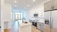 Located at 10410 Sam Furr Road in Huntersville, NC, this thoughtfully designed one-bedroom, one-bathroom residence offers a bright and contemporary living experience with a clean, modern aesthetic throughout. Light-toned hardwood-style flooring flows seamlessly across the entire space, complemented by crisp white walls and recessed lighting that enhance the airy atmosphere.

The open-concept living, dining, and kitchen area serves as the heart of the unit. The kitchen is well-appointed with two-tone cabinetry — white upper cabinets paired with gray lower cabinets — stainless steel appliances including a refrigerator with ice maker, electric range, and microwave, plus a central island with a white quartz countertop and undermount sink with a matte black faucet. The living area is anchored by a ceiling fan and French doors with transom windows that flood the space with natural light and provide access to a private balcony.

The spacious bedroom features a ceiling fan, a large window with outdoor views, and convenient access to two closets, including a walk-in closet outfitted with wire shelving systems. The bathroom offers a dual-sink vanity with gray cabinetry, matte black fixtures, framed mirrors, and a soaking tub with white subway tile surround. Additional utility and in-unit laundry spaces round out this efficiently planned residence.