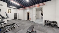 Located at 938 E Fourth St in Bethlehem, Pennsylvania, this fitness center offers a well-equipped workout space within a compact, efficiently designed footprint. The main exercise area features durable patterned carpet tile flooring and an exposed black ceiling with visible ductwork, red-painted piping, and industrial lighting — giving the space a modern, utilitarian aesthetic. A full-length mirrored wall with a light blue cinder block base runs along one side, enhancing the sense of openness and allowing users to monitor their form during workouts. The facility is equipped with a treadmill, elliptical, stationary spin bike, cable machine, adjustable weight bench, and a rack of free weights, making it suitable for a variety of cardio and strength training routines. A wall-mounted flat-screen TV is positioned for viewing during cardio sessions. A standout architectural detail is the set of decorative stained glass windows, hinting at the building's historic character and adding a unique visual contrast to the modern gym interior. Staircase access connects the space to the rest of the building. A private bathroom with wood-look vinyl flooring and clean white walls provides added convenience, along with a closet for storage. The space is rounded out with a fire suppression system, emergency exit signage, and a fire extinguisher, ensuring safety throughout.