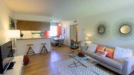 This single-floor residence offers an open-concept layout that seamlessly integrates the living, dining, and kitchen areas into one cohesive, inviting space. The living area is anchored by warm wood-style flooring that extends throughout, complemented by a patterned area rug, a contemporary glass coffee table, and a comfortable sofa accented with vibrant coral and neutral throw pillows. A distinctive sunburst mirror adds a stylish focal point to the main wall.

The dining area sits adjacent to the living space beneath a ceiling fan, providing comfortable air circulation. It features a dining table with upholstered chairs, positioned near a window dressed with bold coral curtains that introduce a lively pop of color and allow natural light to filter in.

The kitchen is partially open to the main living area via a breakfast bar with seating for two, making it ideal for casual dining or entertaining. It is equipped with white appliances, ample cabinetry, and counter space, with teal-accented walls adding a playful contrast to the overall neutral palette.

A hallway visible from the main living area leads to additional rooms, suggesting a well-organized and functional floor plan. The textured ceilings and warm, cohesive color scheme throughout give the space a welcoming and comfortable ambiance.