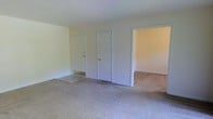 This single-floor residential space offers a clean, straightforward layout designed for comfortable everyday living. The main living area is finished with wall-to-wall carpet in a neutral beige tone, creating a warm and understated foundation throughout the space. The walls are painted in a light, off-white hue that reflects ambient light and contributes to the overall brightness of the interior.

The living area features two interior doors side by side — one leading to an entry or closet space and another providing access to an adjoining room, allowing for a naturally connected flow between spaces. An open doorway to the right leads into a secondary room, also finished with matching carpet, maintaining a consistent aesthetic throughout.

White trim and baseboards frame the doorways and walls, adding a crisp, clean contrast to the neutral palette. The layout is open and uncluttered, offering flexibility in how each area can be arranged and utilized. The even ceiling height and simple architectural lines give the space a calm, no-frills character that is both practical and easy to personalize. Overall, this space presents a modest yet functional single-floor layout well-suited for those seeking a straightforward and livable environment.