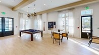 This generously sized single-floor community clubhouse or leasing facility is thoughtfully designed with a contemporary coastal aesthetic that flows seamlessly across its many functional zones. The reception area makes a striking first impression, featuring a wide white quartz counter with wood paneling, navy velvet chairs, globe pendant lighting, and a dramatic sculptural botanical medallion mounted against a geometric lattice backdrop beneath soaring vaulted ceilings with exposed beam detailing. Adjacent to reception, a café-style kitchen area showcases dark cabinetry with brass hardware, open wood shelving displaying curated décor and greenery, herringbone tile backsplash, and a communal dining table beneath a large pendant light. A lounge area provides a warm, residential feel with a fireplace, built-in shelving, sheer floor-to-ceiling drapery, light hardwood flooring, and a chandelier with antler-inspired accents. The game room features a full-size billiards table, pendant lighting, wall-mounted game boards, and abundant natural light from tall windows with sheer curtains. A co-working nook includes a built-in white cabinetry wall with herringbone tile accents and rattan seating. Throughout, the space maintains a bright, airy ambiance unified by a neutral palette and natural textures.
