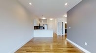 This single-floor space showcases a thoughtfully designed open-concept layout that seamlessly connects the living area to the kitchen. Warm-toned hardwood flooring extends throughout, lending continuity and a welcoming feel to the entire space. The kitchen is outfitted with white cabinetry, a dark brick tile backsplash, stainless steel appliances including a refrigerator and microwave, and a white peninsula that provides both additional prep space and a natural divider between the kitchen and living area. Pendant lighting and flush-mount ceiling fixtures illuminate the kitchen, complementing the recessed lighting found throughout the rest of the space. The walls are painted in a neutral palette, with a contrasting gray accent wall adding a subtle design element. A hallway visible from the main living area leads to additional rooms, including what appears to be a bathroom with dark cabinetry. The overall ambiance is clean, bright, and contemporary, with a minimalist aesthetic that allows the architectural details and finishes to take center stage.