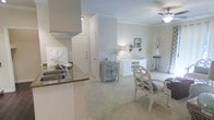 This single-floor residence showcases an open-concept design that connects the kitchen and living areas in one cohesive space. The kitchen is positioned toward the front, featuring a sink with a modern faucet set into dark granite-style countertops, white cabinetry, and a range hood overhead. The layout flows naturally into the main living area, which is finished with plush carpeting and neutral, soft gray-white walls that create a calm, airy atmosphere throughout.

Crown molding runs along the ceiling, adding a refined architectural detail, while a ceiling fan provides both function and a relaxed coastal feel. A dedicated entryway with a solid front door is visible from the main living space, offering a clear sense of the unit's layout.

The living area is styled with coastal-inspired décor, including an ornate white accent chair, a decorative cabinet, and a console table adorned with a starfish motif lamp and wall shelf. The overall ambiance is light, breezy, and welcoming, with a cohesive neutral palette that enhances the sense of openness throughout the space.