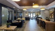 This single-floor space offers a welcoming and well-appointed interior designed with both comfort and functionality in mind. The open layout seamlessly blends a relaxed lounge area with a practical co-working bar station, making it ideal for socializing or focused work. The lounge features plush navy blue and neutral upholstered seating arranged around a rustic wood coffee table, anchored by a textured area rug over light wood-plank flooring that runs throughout the space. A standout design element is the decorative use of birch tree trunks, which serve as natural vertical accents and bring an organic, nature-inspired aesthetic to the interior. Overhead, large drum pendant lights with warm amber shades hang from a rich brown-painted ceiling section, creating a cozy and inviting ambiance. Recessed lighting supplements the pendants to ensure even illumination. Along one side of the space, a long bar-height wooden countertop with black stools provides a functional workspace or casual dining area. Large windows allow natural light to filter in, offering views of the greenery outside. A secondary area, partially visible through glass partitions, suggests additional flexible space for meetings or private use.