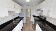This single-story space showcases a thoughtfully designed galley-style kitchen as its centerpiece. The kitchen features crisp white shaker-style cabinetry with brushed nickel hardware lining both walls, providing generous storage throughout. Dark granite countertops offer a striking contrast against the bright cabinetry, and are present on both sides of the kitchen corridor. On the left side, a stainless steel undermount sink with a modern pull-down faucet is paired with a dishwasher and additional cabinet storage. The right side is equipped with a stainless steel electric range topped with a ventilation hood, ideal for everyday cooking. A stainless steel refrigerator anchors the far end of the kitchen, beyond which a hallway leads to additional rooms. The flooring throughout is a warm-toned luxury vinyl plank, adding durability and visual continuity. A flush-mount ceiling light illuminates the space, complemented by a textured ceiling. The overall layout is efficient and functional, maximizing every inch of available space while maintaining a clean, modern aesthetic.