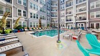 Located at 1325 Boland Pl in Richmond Heights, Missouri, this dynamic outdoor amenity courtyard is nestled within a multi-story residential community, creating an inviting, enclosed retreat surrounded by balconied residential floors on all sides. At the heart of the space is a generously sized resort-style swimming pool, framed by a spacious concrete pool deck lined with modern lounge chairs accented in bold orange and white, ideal for relaxing in the sun. Overhead patio heaters extend the usability of the space across seasons. Adjacent to the pool, a dedicated lounge area features a central fire pit surrounded by colorful teal Adirondack chairs and additional seating arrangements, offering a comfortable social gathering spot. A gas grill station with granite countertops provides an outdoor cooking area, conveniently positioned near a bold, geometric mural in teal, orange, and gray that serves as a striking focal point and adds an artistic, energetic character to the courtyard. The ground-level terrace also includes shaded seating nooks with stylish lounge chairs and planters with greenery, softening the hardscape. The multi-story building façade, clad in gray lap siding with black-railed balconies, encloses the courtyard and creates a sense of privacy and community while flooding the space with natural light throughout the day.