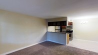 This single-floor residence offers a clean, open layout that combines the living area and kitchen in a practical and cohesive design. The living space features wall-to-wall carpeting in a neutral tone, creating a comfortable and inviting atmosphere, while smooth walls painted in a soft beige provide a warm, understated backdrop throughout.

The kitchen is defined by a raised peninsula counter with dark granite-style countertops, which serves as a natural divider between the living and cooking areas. Dark wood cabinetry — including upper cabinets with glass-panel doors — adds depth and contrast to the space. The kitchen is outfitted with stainless steel appliances, including a refrigerator, a range with oven, and an over-the-range microwave, offering a modern and functional cooking environment. Light-toned flooring in the kitchen contrasts neatly with the carpeted living area, visually distinguishing the two zones.

The space benefits from a straightforward, efficient floor plan that maximizes usability. Clean white baseboards frame the rooms, and recessed lighting in the kitchen area adds to the functional illumination. Overall, the unit presents a tidy, move-in ready interior with a cohesive neutral palette suitable for a variety of living styles.
