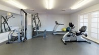 This single-level fitness room offers a comprehensive workout environment designed to accommodate a variety of exercise routines. The space is outfitted with durable rubber flooring throughout, providing both safety and sound absorption during training sessions. A large wall-mounted mirror on one end of the room enhances the sense of space while allowing users to monitor their form during exercises.

The room is equipped with a multi-function cable weight machine, a treadmill, a recumbent stationary bike, and an ab training machine, providing options for both strength and cardiovascular training. A dedicated weight rack holding a selection of medicine balls and dumbbells adds further versatility. Exercise balls in green and yellow are also available for core and flexibility work.

Natural light enters the room through two sets of French doors, each fitted with multi-pane glass, offering views of the outdoor surroundings and providing convenient access to an exterior patio or courtyard. Overhead fluorescent lighting ensures consistent illumination throughout the space. The textured ceiling and neutral white walls give the room a clean, functional aesthetic suited to an active fitness environment.