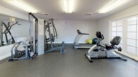 This single-level fitness room offers a comprehensive workout environment designed to accommodate a variety of exercise routines. The space is outfitted with durable rubber flooring throughout, providing both safety and sound absorption during training sessions. A large wall-mounted mirror on one end of the room enhances the sense of space while allowing users to monitor their form during exercises.

The room is equipped with a multi-function cable weight machine, a treadmill, a recumbent stationary bike, and an ab training machine, providing options for both strength and cardiovascular training. A dedicated weight rack holding a selection of medicine balls and dumbbells adds further versatility. Exercise balls in green and yellow are also available for core and flexibility work.

Natural light enters the room through two sets of French doors, each fitted with multi-pane glass, offering views of the outdoor surroundings and providing convenient access to an exterior patio or courtyard. Overhead fluorescent lighting ensures consistent illumination throughout the space. The textured ceiling and neutral white walls give the room a clean, functional aesthetic suited to an active fitness environment.