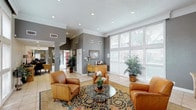 This single-floor space functions as a generously sized communal lounge or reception area, thoughtfully designed to balance comfort and functionality. The room is finished with light-toned ceramic tile flooring throughout, complemented by a richly patterned area rug that anchors the central seating arrangement of tan leather armchairs surrounding a glass-top coffee table.

A standout architectural feature is the substantial brick fireplace fitted with a crisp white mantle, decorated with framed artwork and candle accents, lending the space a warm, classic character. Adjacent to the fireplace, a wall-mounted flat-screen television adds a modern, practical element.

Large multi-panel windows with white horizontal blinds line the walls, allowing abundant natural light to fill the interior while offering views of the surrounding greenery outdoors. The walls are finished in a warm gray tone, accented by white crown molding and recessed lighting embedded in the textured ceiling, which adds depth and dimension overhead.

A wooden console table with decorative accessories and a coffee station provides additional functionality along one wall. Potted indoor plants are positioned throughout, enhancing the welcoming ambiance. An open corridor connects this lounge to adjacent areas, suggesting a well-organized, flowing floor plan designed for ease of movement and accessibility.