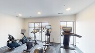 This fitness center is a thoughtfully designed, single-floor workout facility that offers a comprehensive range of exercise options in a clean, modern environment. The cardio area is well-stocked with multiple stationary bikes, an elliptical trainer, and a treadmill, each equipped with individual digital screens for an engaging workout experience. Large sliding glass doors and wide windows flood the space with natural light and open to an outdoor terrace, providing expansive views that enhance the exercise environment. The strength training area features a full rack of hex dumbbells in graduated weights, set against a warm, textured accent wall with a geometric pattern that adds a stylish visual anchor to the space. Built-in shelving and cabinetry in natural wood tones offer convenient storage for towels and accessories, complemented by a sturdy wooden bench for seating. A wall-mounted flat-screen television is positioned for easy viewing throughout the space. The flooring consists of durable, patterned carpet tiles in muted blue-grey tones, providing comfort and stability underfoot. Recessed and flush-mount ceiling lighting ensures the entire space is evenly and adequately illuminated. The overall ambiance is contemporary and functional, designed to support a focused and motivating fitness experience.