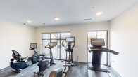 This fitness center is a thoughtfully designed, single-floor workout facility that offers a comprehensive range of exercise options in a clean, modern environment. The cardio area is well-stocked with multiple stationary bikes, an elliptical trainer, and a treadmill, each equipped with individual digital screens for an engaging workout experience. Large sliding glass doors and wide windows flood the space with natural light and open to an outdoor terrace, providing expansive views that enhance the exercise environment. The strength training area features a full rack of hex dumbbells in graduated weights, set against a warm, textured accent wall with a geometric pattern that adds a stylish visual anchor to the space. Built-in shelving and cabinetry in natural wood tones offer convenient storage for towels and accessories, complemented by a sturdy wooden bench for seating. A wall-mounted flat-screen television is positioned for easy viewing throughout the space. The flooring consists of durable, patterned carpet tiles in muted blue-grey tones, providing comfort and stability underfoot. Recessed and flush-mount ceiling lighting ensures the entire space is evenly and adequately illuminated. The overall ambiance is contemporary and functional, designed to support a focused and motivating fitness experience.