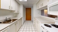 This single-floor unit offers a practical and functional layout spread across several well-defined living spaces. The carpeted living and bedroom areas are finished in neutral beige tones, creating a calm and cohesive atmosphere throughout. Multiple sliding glass doors fitted with vertical blinds allow natural light to fill the rooms while providing direct access to an outdoor patio area, where brick surroundings are visible. Baseboard heating runs consistently along the walls, ensuring efficient climate control throughout. A ceiling fan with a light fixture is installed in the main living area, adding both ventilation and overhead illumination.

The galley-style kitchen features white cabinetry lining both walls, light countertops, a tile floor, and a drop ceiling with flush lighting. A range and sink are positioned within the kitchen's efficient corridor layout, with ample cabinet storage above and below.

The primary bedroom includes a wide double-door closet and access to a full bathroom. One bathroom features accessibility-focused grab bars near the toilet and a tub-shower combination, while a second bathroom offers a compact vanity, mirrored storage, and a tiled shower. Both bathrooms are finished with white tile flooring and light-toned cabinetry.