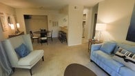 This single-floor residence showcases an open-concept layout that seamlessly connects the living, dining, and kitchen areas into one cohesive space. The living area is furnished with comfortable seating and features warm, neutral-toned carpeting that extends throughout, creating a cozy and inviting atmosphere. A stylish floor lamp adds ambient lighting, complementing the overall warmth of the space.

The dining area accommodates a round table with seating for multiple guests, positioned adjacent to the kitchen for easy entertaining. A half-wall partition with a breakfast bar counter separates the kitchen from the main living and dining areas, offering bar-stool seating for casual dining. This architectural detail adds visual interest while maintaining the open feel of the floor plan.

The kitchen is accessible beyond the partition and features cabinetry providing ample storage. Overhead HVAC vents are integrated into the ceiling, ensuring year-round comfort throughout the space. Neutral-toned walls and light-colored flooring in the kitchen area keep the space feeling bright and clean.

The overall design is practical and well-organized, with a neutral color palette of beige, cream, and soft blue accents that create a calm and welcoming environment. The open flow between rooms makes the space feel spacious and easy to navigate.