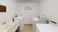 This shared laundry facility is designed with functionality and cleanliness in mind. The single-floor space is finished with bright white walls and durable speckled linoleum flooring, creating a tidy, well-lit environment for residents. Multiple commercial-grade Maytag top-load washing machines and matching dryers are arranged along the walls, providing ample capacity for concurrent use. Each machine is equipped with a WASH coin-operated payment system, offering convenient and straightforward operation. A deep utility sink is positioned along one wall, ideal for hand-washing, pre-treating garments, or other laundry-related tasks. A cork bulletin board is mounted on the wall, serving as a practical communication hub for building notices or community information. A charming laundry-themed word art decal adorns one wall, adding a lighthearted touch to the otherwise utilitarian space. The room is accessed through a dark green door with a frosted glass panel and an automatic door closer, ensuring privacy and ease of entry. Overall, the space is thoughtfully laid out to maximize usability while maintaining a clean and organized atmosphere.