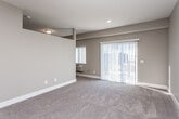 This single-floor unit offers a practical and well-designed layout, combining comfort with modern finishes throughout. The open-concept living area is finished with plush wall-to-wall carpeting and neutral taupe-toned walls, creating a warm and understated ambiance. Architectural details include angled ceiling lines and a raised soffit that add visual interest to the space. A wall-mounted HVAC unit provides independent climate control, while a side window allows natural light to filter in.

The living area opens toward a peninsula-style kitchen, which is separated by a breakfast bar with granite-style countertops. The kitchen is equipped with stainless steel appliances, including a refrigerator, electric range, and over-the-range microwave. Crisp white shaker-style cabinetry lines the walls, complemented by wood-look luxury vinyl plank flooring that extends throughout the kitchen and adjacent areas. Pendant lighting above the peninsula adds a refined touch.

A set of sliding glass doors, dressed with vertical blinds, connects the living area to an outdoor space, bringing in additional natural light. The bathroom features a full tub-and-shower combination, a white vanity with granite-style countertops, and wood-look flooring, completing the unit's cohesive aesthetic.