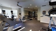 This fitness room is designed to support a comprehensive workout routine in a clean, functional setting. The space is finished with dark rubber flooring that provides durability and cushioning underfoot, while neutral gray walls and white trim create a crisp, modern aesthetic. Recessed lighting throughout the ceiling ensures even illumination across the room, complemented by two ceiling fans that promote air circulation during workouts. A large wall mirror along one side visually expands the space and allows users to monitor their form. The equipment layout is well-organized, with cardio machines — including an elliptical, stationary bike, and treadmill — positioned toward the perimeter. A multi-tiered dumbbell rack offers a range of free weights, and a cable machine system provides options for strength and resistance training. A wall-mounted screen near the entrance offers interactive or informational content for users. Natural light filters in through a window on one side, adding brightness to the space. Overall, the room is thoughtfully arranged to maximize usable floor space while offering a well-rounded selection of fitness equipment.