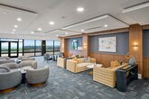 This expansive rooftop lounge offers a refined, upscale environment designed for both social gatherings and private events. The open floor plan is anchored by a central main lounge area with multiple distinct seating arrangements, featuring plush curved sofas, accent chairs, and structured leather seating grouped around stylish side tables. Warm wood-paneled walls, coffered ceilings with recessed lighting, and patterned carpet tile flooring throughout create a cohesive and polished aesthetic.

A dedicated bar area stretches along one side of the space, with dual-sided white quartz countertops, wood cabinetry with brass hardware, terrazzo flooring, and pendant lighting overhead. High-backed bar stools line both counters, making it ideal for serving large groups. Floor-to-ceiling windows in multiple zones frame dramatic panoramic city skyline views, while a glass-walled private meeting or event room provides a flexible enclosed space. A separate lounge alcove along the perimeter offers intimate seating directly adjacent to the outdoor terrace, which is accessible via large sliding glass doors. Curated artwork, circular mirrors, gold-toned sconces, and thoughtfully placed accent lighting add warmth and character throughout, completing this sophisticated elevated venue.