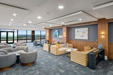 This expansive rooftop lounge offers a refined, upscale environment designed for both social gatherings and private events. The open floor plan is anchored by a central main lounge area with multiple distinct seating arrangements, featuring plush curved sofas, accent chairs, and structured leather seating grouped around stylish side tables. Warm wood-paneled walls, coffered ceilings with recessed lighting, and patterned carpet tile flooring throughout create a cohesive and polished aesthetic.

A dedicated bar area stretches along one side of the space, with dual-sided white quartz countertops, wood cabinetry with brass hardware, terrazzo flooring, and pendant lighting overhead. High-backed bar stools line both counters, making it ideal for serving large groups. Floor-to-ceiling windows in multiple zones frame dramatic panoramic city skyline views, while a glass-walled private meeting or event room provides a flexible enclosed space. A separate lounge alcove along the perimeter offers intimate seating directly adjacent to the outdoor terrace, which is accessible via large sliding glass doors. Curated artwork, circular mirrors, gold-toned sconces, and thoughtfully placed accent lighting add warmth and character throughout, completing this sophisticated elevated venue.