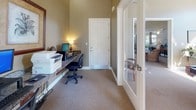 This single-floor business center is designed to provide a functional and comfortable workspace for its users. The room is finished with warm taupe-toned walls and neutral wall-to-wall carpeting, creating a professional yet inviting atmosphere throughout.

A continuous granite-topped countertop runs along the length of the main wall, offering generous workspace for multiple users simultaneously. The workstations are equipped with desktop computers, keyboards, and task lighting provided by classic table lamps that add a warm ambient glow. A multifunction printer is also integrated into the workspace, ensuring full office functionality. A corkboard mounted on the wall serves as an informational display area, adding a practical organizational element to the room.

Natural light is a defining feature of this space. A full-length glass-paned exterior door with a transom window above it floods the room with sunlight, offering views of the surrounding outdoor landscape. This creates a bright and airy environment that enhances productivity. A second interior glass-paned door connects this business center to an adjacent space, further contributing to the open and accessible feel.

The elevated ceiling height adds a sense of spaciousness to the room. Ergonomic rolling office chairs are positioned throughout, accommodating several workstation users at once. The overall design balances practicality with comfort, making this business center a well-equipped and welcoming environment for day-to-day productivity needs.
