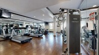 Located at 3250 Market St in Riverside, California, this fitness center offers a well-appointed exercise room designed to support a comprehensive workout experience. The space features rich hardwood flooring throughout, which lends a polished and professional ambiance to the room. Recessed lighting illuminates the area evenly, while large floor-to-ceiling mirrors line the walls, visually expanding the space and allowing users to monitor their form during exercise. Natural light filters in through several windows, with sliding glass doors providing access to an outdoor balcony area and views of the surrounding neighborhood. The cardio zone is equipped with multiple treadmills, elliptical machines, and a stationary bike, while the strength training area includes a Nautilus Instinct cable machine, a dumbbell rack, and an adjustable weight bench. A rowing machine and stability ball round out the functional fitness offerings. Structural columns are integrated into the layout, and dark wood crown molding adds a refined architectural detail to the white walls and ceiling. A posted fitness center rules sign indicates a managed, community-oriented facility. The overall atmosphere is clean, functional, and thoughtfully outfitted for a range of fitness goals.