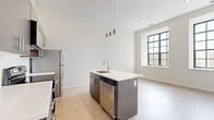 This thoughtfully designed one-bedroom, one-bathroom unit offers a seamless blend of modern aesthetics and functional living across a single floor. The open-concept kitchen and living area serves as the heart of the home, anchored by a sleek kitchen island with a white quartz countertop, an undermount sink, and stainless steel appliances including a refrigerator, range, microwave, and dishwasher. Dark wood-grain cabinetry extends to the upper walls, complemented by stylish pendant lighting that hangs from the dramatically tall ceilings, amplifying the sense of openness. Light-toned hardwood flooring flows throughout the living areas, adding warmth to the contemporary palette. Large, bold steel-framed windows flood both the living space and bedroom with abundant natural light while framing urban views of the surrounding cityscape. The bedroom is generously proportioned, finished with plush carpet, soaring ceilings, and a walk-in closet for ample storage. A light-filled hallway connects the living areas to the private quarters. The bathroom features large-format tile flooring, a modern floating vanity with a white countertop, and a spacious walk-in shower enclosed by a sleek sliding glass door. Clean lines and a neutral color palette lend the entire unit a bright, contemporary ambiance.