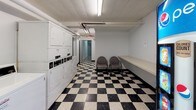 This shared amenity space is designed to serve the practical needs of its occupants, combining a fully equipped laundry room with a vending and lounge area. The entire space is finished with a striking black-and-white checkered tile floor that runs throughout, lending a retro, utilitarian character to the interior.

The laundry area houses a row of coin-operated top-load washing machines and stacked dryers, providing ample capacity for multiple users. A built-in wall-mounted counter runs along one side, offering a convenient surface for folding clothes or sorting laundry. Exposed overhead piping and fluorescent tube lighting give the space a functional, no-frills aesthetic, while white walls keep the environment clean and bright.

Adjacent to the laundry room, the vending and lounge area features a Pepsi beverage vending machine alongside trash receptacles and an electrical panel mounted on the wall. A small seating area with upholstered chairs provides a comfortable waiting space. Additional wall-mounted counters offer further utility. Fluorescent overhead lighting illuminates the entire area, and a corridor connects the two zones, creating a cohesive, practical shared amenity space.
