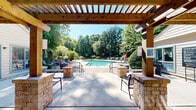 Located at 1507 Pine Dr in Atlanta, Georgia, this outdoor pool facility offers a well-appointed aquatic retreat set against a lush backdrop of mature pine and hardwood trees. The centerpiece is a generously sized swimming pool with a distinctive geometric shape, featuring both shallow and deeper sections clearly marked for safety. The crystal-clear, blue-green water is complemented by a wide surrounding deck finished in warm-toned pavers, providing ample space for lounging and relaxation.

A handsome wooden pergola with a ceiling fan anchors one end of the pool area, framing views of the water and greenery beyond. Brick columns and built-in seating areas add architectural character to the covered space, creating a shaded gathering spot ideal for warm Georgia days. The pool deck is furnished with lounge chairs and tables topped with vibrant orange umbrellas, offering both sun and shade options throughout the day.

The pool area is fully enclosed by a black metal fence, ensuring privacy and security. Lush landscaping, including trimmed shrubs and ornamental plantings, lines the perimeter, while towering trees provide a natural canopy that enhances the sense of seclusion. A clubhouse-style building is situated adjacent to the pool, adding functional support to this outdoor amenity. The overall ambiance is serene and resort-like, making it a standout outdoor feature of this Atlanta property.