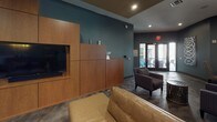 This sophisticated single-floor community lounge is designed with a contemporary aesthetic, combining dark, moody tones with warm wood accents and polished concrete flooring throughout. The open-concept layout accommodates multiple distinct activity zones, each thoughtfully curated with upscale furnishings and curated artwork adorning the walls.

A billiards table anchors one section of the space, surrounded by casual seating that encourages social gathering. Nearby, high-top bar tables with dark wood and metal bases offer a comfortable spot for conversation, positioned just steps from floor-to-ceiling glass sliding doors that flood the area with natural light and frame views of the outdoor pool and courtyard beyond.

A dedicated media nook features a built-in wood-paneled entertainment wall with a mounted flat-screen television, complemented by a plush sofa and textured tile accent wall, creating an intimate retreat within the larger space. Additional lounge seating areas are arranged throughout, incorporating leather sofas, upholstered armchairs, and accent tables.

Rich teal and charcoal wall colors set a bold, modern tone, while decorative wall sculptures and large-scale abstract paintings add artistic character. Direct exterior access connects seamlessly to the outdoor amenity area, making this a versatile and inviting hub for relaxation and entertainment.