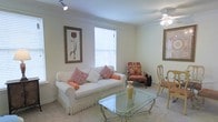 This single-floor space features a well-appointed living area designed with a classic, traditional aesthetic. The room is finished with light neutral walls and carpeted flooring, creating a warm and inviting atmosphere throughout. A ceiling fan with a light fixture is centrally positioned overhead, providing both air circulation and ambient lighting.

The living area is anchored by a white upholstered sofa adorned with coral and floral-patterned accent pillows, complemented by a matching fringe detail along its base. A glass-top coffee table with an ornate metal frame sits in front of the sofa, adding an elegant touch to the space. A coordinating floral armchair with carved wooden legs provides additional seating nearby.

Natural light enters through a large window fitted with horizontal blinds, brightening the room considerably. Decorative framed botanical and ornamental artwork on the walls adds personality and a refined charm. A dining area is partially visible, featuring wooden chairs with upholstered seats, suggesting a seamlessly connected layout. Overall, the space presents a traditional, well-curated interior with consistent warm tones and thoughtful decorative touches.