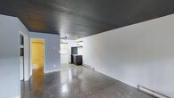 Located at 917 Dacian Avenue in Durham, North Carolina, this single-floor space presents a distinctive industrial-modern aesthetic that sets it apart. The interior is immediately striking, with polished concrete floors that reflect light throughout the open layout, creating a sleek and durable foundation underfoot. The bold, dark textured ceiling adds dramatic contrast against the light gray walls, lending the space a contemporary, edgy character.

The open floor plan allows for flexible use of the space, with a kitchen area positioned toward the rear. The kitchen is equipped with a refrigerator and range, complemented by a wall-mounted shelf for additional storage. A baseboard heater runs along the wall, ensuring comfort year-round. Natural light filters in through a window near the kitchen, brightening the area and providing ventilation.

A vibrant yellow door adds a pop of color and serves as a distinctive architectural accent, contributing to the space's eclectic personality. The combination of raw, industrial materials — polished concrete, dark ceilings — with clean white and gray walls creates a balanced and functional environment. This space is well-suited for those who appreciate minimalist design with bold, expressive details in the heart of Durham.
