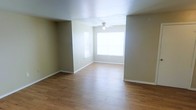 This single-floor space presents a straightforward and practical layout designed for comfortable, everyday living. The main living area is immediately welcoming, featuring warm hardwood-style flooring that extends throughout and adds a sense of continuity and warmth to the space. Neutral beige-toned walls provide a versatile backdrop, making it easy to personalize with a variety of décor styles.

Natural light plays a key role in the ambiance of the living area, with a large picture window drawing in generous amounts of daylight and creating a bright, open atmosphere. A ceiling fan centered in the room enhances air circulation and contributes to the overall comfort of the space. Clean white baseboards frame the flooring neatly, adding a crisp, finished look to the interior.

The open feel of the main room, defined by a subtle architectural division, allows for flexible furniture arrangements and functional use of the available space. The overall design is clean and understated, prioritizing practicality and livability. With its neutral palette, natural light, and quality flooring, this space offers a solid foundation for a comfortable and well-organized living environment.