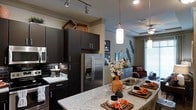 This single-floor residence offers a thoughtfully designed open-concept layout that seamlessly connects the kitchen and living areas, creating a cohesive and inviting atmosphere. The kitchen is a standout feature, showcasing sleek dark cabinetry that extends to the upper walls, granite countertops, and a suite of stainless steel appliances, including a French door refrigerator with an in-door ice and water dispenser. A dark subway tile backsplash adds a sophisticated contrast to the lighter countertops, while pendant lighting over the island and track lighting overhead ensure the space is well-lit and functional. The kitchen island provides generous prep and dining space, making it ideal for both everyday use and entertaining. Crown molding details frame the ceilings throughout, adding a refined architectural touch. The adjacent living area benefits from natural light streaming in through large windows dressed with neutral drapes. A ceiling fan with a light fixture enhances comfort in the living space. The overall ambiance is modern and upscale, with a cohesive dark-toned palette balanced by lighter walls and countertops, resulting in a space that feels both elegant and welcoming.