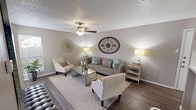 Located in Fort Worth, Texas, this well-appointed single-floor residence at 4858 River Ranch Blvd offers a cohesive and inviting interior defined by warm neutral tones, wood-look flooring, and tasteful decorative accents throughout. The living area serves as the heart of the home, featuring a ceiling fan, textured ceiling, and generous natural light filtering through vertical blinds. Comfortable seating, a mirrored glass coffee table, and decorative wall art create a welcoming and stylish atmosphere. A large bold canvas and sunburst wall medallions add personality to the space. The kitchen is thoughtfully designed with crisp white cabinetry, light countertops, a mosaic tile backsplash, and black appliances, including a dishwasher and range. Track lighting overhead ensures a bright and functional workspace. The bedroom is a serene retreat, with soft neutral walls, a textured ceiling, rich hardwood-style flooring, and a large window dressed with sheer curtains that allow natural light to filter in gently. A dedicated vanity nook adjacent to the bathroom offers an intimate space with tufted seating, abstract wall art, and a sleek console table. The bathroom features white cabinetry and a well-lit vanity mirror. Every detail of this home reflects a refined, contemporary sensibility.
