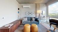 This single-floor space showcases a thoughtfully designed interior that blends industrial and contemporary aesthetics. Exposed concrete ceilings throughout lend an urban character, while light hardwood flooring provides a warm contrast and unifies the living areas.

The living area is anchored by a comfortable gray sofa accented with decorative pillows, complemented by a walnut media credenza and a pair of tan leather ottomans. Built-in open shelving offers practical storage and display space, while a wall-mounted mini-split system ensures climate control. Framed artwork adds a curated touch to the space.

The kitchen features full-height, flat-front cabinetry in a light wood finish, stainless steel appliances including a gas range, oven, dishwasher, and microwave, along with a clean white countertop and a single-basin sink. Large windows flood the kitchen with natural light and offer open outdoor views.

The bathroom is finished in a minimalist style with light wood vanity cabinetry, a white countertop, a large mirror, and a combined tub and shower. A patterned accent rug adds warmth to the otherwise neutral palette. The floor plan also reveals in-unit laundry, providing added convenience throughout the well-organized layout.