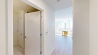 Located in Baltimore, MD 21231, this one-bedroom, one-bathroom unit is thoughtfully designed to maximize every square foot of its compact footprint. The open layout flows from a welcoming hallway into the main living areas, all finished with light wood flooring that enhances the bright, airy atmosphere throughout.

The kitchen is a standout feature, outfitted with stainless steel appliances including a refrigerator, range, microwave, and dishwasher. White upper cabinetry contrasts beautifully with warm wood lower cabinets, while a soft green subway tile backsplash and white countertops add a modern, clean aesthetic. A movable island provides additional prep and dining space.

The living area benefits from large floor-to-ceiling windows that flood the space with natural light and offer sweeping views of the Baltimore cityscape. The bedroom is a comfortable, well-proportioned room with a built-in closet and additional closet storage available in the hallway, offering practical organization solutions.

The bathroom features a streamlined vanity with warm wood cabinetry, white countertops, a large mirror, and clean, neutral finishes. The overall design of the unit is modern and minimalist, with crisp white walls and efficient use of space making it a functional and stylish urban residence.