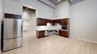 This single-floor unit offers a well-organized layout comprising a kitchen, a full bathroom, and three hallway areas. The kitchen is a standout feature, designed in an L-shaped configuration with rich dark-wood cabinetry extending to impressive heights, white countertops, and a full suite of stainless steel appliances including a wall oven, dishwasher, refrigerator, and a built-in cooktop with range hood. The open layout of the kitchen area benefits from soaring ceilings with exposed ductwork and structural beams, lending an industrial loft aesthetic to the space. Light hardwood flooring flows throughout the main areas, complementing the neutral gray walls and creating a bright, airy feel. A separate carpeted nook provides a softer, quieter zone within the unit. The bathroom is clean and functional, featuring white subway tile surrounding a full soaker tub and shower combo with grab bars, a floating vanity with a white countertop, a framed mirror, and light tile flooring. The hallway areas connect the rooms efficiently, with one hallway visible near the entry door. A wall-mounted mini-split system provides climate control, and recessed lighting throughout ensures consistent illumination across all areas of the unit.