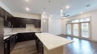 This single-floor residence showcases a contemporary open-concept design that seamlessly connects the kitchen, dining, and living areas. The kitchen is a standout feature, appointed with dark espresso shaker cabinets, bright white quartz countertops, and a full stainless steel appliance suite including a French door refrigerator, range, microwave, and dishwasher. A textured stone tile backsplash adds visual depth, while a generously sized center island with pendant lighting overhead serves as an ideal gathering and prep space. Dark hardwood-style flooring flows throughout the main living areas, complemented by recessed lighting and a ceiling fan in the living room. A large window with blinds allows natural light to fill the space, offering an open, airy feel. The bedrooms are finished with plush carpet and feature windows providing additional natural light. The bathroom is well-appointed with a dual-sink vanity featuring dark cabinetry, white quartz counters, a large framed mirror, and a tiled tub-shower combination with decorative tile accents and light porcelain tile flooring throughout.