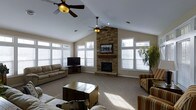 This single-floor living room is defined by its vaulted ceilings, which lend a sense of openness and grandeur to the space. Twin ceiling fans are strategically positioned along the ceiling's peak, ensuring comfortable airflow throughout the room. The focal point of the space is a striking floor-to-ceiling stacked stone fireplace, which anchors the room with a rustic yet refined aesthetic. A wooden mantel shelf sits above the firebox, and a flat-screen television is mounted directly on the stone surround, blending modern convenience with classic design. Flanking the fireplace, multiple large windows with white trim allow generous amounts of natural light to flood the interior, brightening the neutral-toned carpet and warm beige walls. The seating arrangement includes a tan sofa, a striped armchair, and additional casual seating, all oriented around a distinctive trunk-style coffee table that adds a unique, characterful touch. A secondary television and media console occupy one corner, offering additional entertainment options. A lush indoor plant near the windows adds a natural, organic element to the overall warm and inviting ambiance.