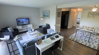 This thoughtfully designed single-floor residence offers an open-concept layout that seamlessly connects the living, dining, and kitchen areas, creating a spacious and cohesive environment. The living area is anchored by a comfortable sectional sofa and complementary armchair, arranged around a round white coffee table atop a patterned area rug. A media console with a flat-screen TV and built-in shelving provides both function and visual interest along one wall, while a framed landscape artwork adds a refined touch above the seating area. A sleek white desk with ample workspace serves as a dedicated work-from-home zone, positioned to take advantage of the open flow of the room. Rich hardwood-style flooring runs throughout, grounding the neutral gray and white color palette and lending warmth to the overall ambiance. The kitchen is visible from the main living space and features a full-size refrigerator, contributing to the home's practical design. A dining area with a table and chairs sits adjacent to the kitchen, ideal for casual meals or entertaining. The interiors are bathed in soft, warm lighting from a combination of floor lamps and overhead fixtures, creating an inviting and comfortable atmosphere throughout.