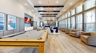 Located at Avida Village Circle in Orlando, Florida, this single-floor community clubroom offers a generously sized, open-plan gathering space designed with a contemporary aesthetic. The room is anchored by soaring ceilings with striking dark exposed beams and illuminated by a combination of recessed lighting and a mid-century modern sputnik chandelier, creating a warm yet sophisticated ambiance throughout.

The space is thoughtfully zoned for multiple activities. A full-size pool table serves as a centerpiece for the game area, positioned near a wall-mounted fireplace display and a large flat-screen TV. Adjacent to this, a substantial wood-and-steel dining table surrounded by tan leather chairs provides a versatile gathering spot for meals or group activities.

A lounge area features a large navy sectional sofa, neutral accent chairs, and a concrete coffee table, all set against a floor-to-ceiling bank of windows that floods the space with natural light and offers views of the outdoor surroundings. Additional seating nooks with plush armchairs line the windowed wall, ideal for quieter moments.

The kitchenette is outfitted with white shaker cabinetry, light stone countertops, a stainless steel refrigerator, microwave, and a striking metallic tile backsplash with open shelving. Light wood-look flooring unifies the entire space, complementing its modern, inviting character.