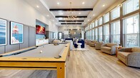 Located at Avida Village Circle in Orlando, Florida, this single-floor community clubroom offers a generously sized, open-plan gathering space designed with a contemporary aesthetic. The room is anchored by soaring ceilings with striking dark exposed beams and illuminated by a combination of recessed lighting and a mid-century modern sputnik chandelier, creating a warm yet sophisticated ambiance throughout.

The space is thoughtfully zoned for multiple activities. A full-size pool table serves as a centerpiece for the game area, positioned near a wall-mounted fireplace display and a large flat-screen TV. Adjacent to this, a substantial wood-and-steel dining table surrounded by tan leather chairs provides a versatile gathering spot for meals or group activities.

A lounge area features a large navy sectional sofa, neutral accent chairs, and a concrete coffee table, all set against a floor-to-ceiling bank of windows that floods the space with natural light and offers views of the outdoor surroundings. Additional seating nooks with plush armchairs line the windowed wall, ideal for quieter moments.

The kitchenette is outfitted with white shaker cabinetry, light stone countertops, a stainless steel refrigerator, microwave, and a striking metallic tile backsplash with open shelving. Light wood-look flooring unifies the entire space, complementing its modern, inviting character.