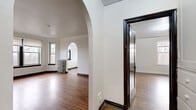 This single-floor residence blends classic architectural details with functional living spaces throughout. The layout is defined by elegant arched doorways that connect rooms seamlessly, creating a sense of openness and architectural character. Warm hardwood flooring extends through the primary living and bedroom areas, complemented by dark-trimmed baseboards and door frames that add a rich, traditional contrast to the white walls.

The spacious living area features a sweeping bay window arrangement that invites an abundance of natural light and outdoor views, while a cast-iron radiator underscores the space's vintage charm. Crown molding runs throughout, adding a refined finishing touch to the ceilings.

The bedrooms are well-proportioned, each equipped with a window for natural light, radiator heating, and closet storage. The richly stained wooden doors throughout the unit reinforce the classic aesthetic.

The bathroom is a standout feature, finished with green ceramic wall tiles surrounding the bathtub and complementary yellow-green floor tiles, offering a distinctive, retro character. The kitchen includes wood cabinetry, a stainless steel sink, tile backsplash, and full-size refrigerator, laid over durable tile flooring. An additional tiled room with multiple windows provides versatile flex space. Overall, the residence offers a cohesive blend of period details and practical amenities.