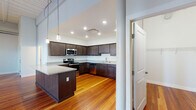 This single-floor space blends modern updates with classic architectural character throughout. Rich, warm-toned hardwood floors run continuously through the living areas, bedrooms, and kitchen, lending a cohesive and timeless feel to the entire unit.

The kitchen is a standout feature, offering generous cabinetry in a deep espresso finish, granite countertops, and a full suite of stainless steel appliances including a range, microwave, dishwasher, and refrigerator. A central island provides additional prep and counter space, while pendant lighting adds a refined touch overhead. Recessed lighting ensures the kitchen remains bright and functional.

The main living area impresses with its open layout, exposed ductwork, and a distinctive wood-paneled ceiling that speaks to the building's character. Multiple windows invite natural light and contribute to the airy, loft-like ambiance.

The bathroom is designed with accessibility in mind, featuring a roll-in shower with grab bars, a comfort-height toilet with safety rails, classic mosaic tile flooring, and a granite-topped vanity. A dedicated laundry room with a stacked washer and dryer adds everyday convenience.

Bedrooms are well-proportioned, with hardwood floors, natural light from grid-pane windows, and a spacious walk-in closet equipped with wire shelving for organized storage.