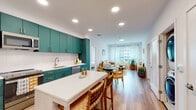 Located at 3901 O'Donnell Street in Baltimore's vibrant Canton neighborhood, this single-floor residence offers a thoughtfully designed open-concept layout that seamlessly connects the kitchen, dining, and living areas. The kitchen is a standout feature, showcasing bold teal flat-front cabinetry with brushed gold hardware, a white subway tile backsplash, a white quartz waterfall island with bar seating, and a full suite of stainless steel appliances. Light wood-tone flooring runs throughout the main living areas, enhancing the bright, airy atmosphere created by recessed lighting and large windows dressed in sheer curtains.

The living room benefits from generous natural light and urban views, providing a welcoming, relaxed ambiance. A secondary room with soft sage-toned walls and natural light serves well as a bedroom or flexible-use space. The primary bedroom features soft carpet, corner windows, and access to a spacious walk-in closet outfitted with built-in shelving and hanging rods on both sides. The bathroom is finished with classic black-and-white hexagonal floor tile, sage green wall tile in the tub surround, and gold fixtures, adding a refined, retro-modern touch to the overall design.