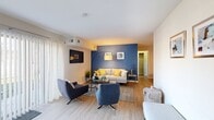This single-floor residence offers a thoughtfully arranged layout that balances comfort and functionality across its main living areas. The living room serves as the centerpiece of the home, featuring light hardwood-style flooring, a bold navy blue accent wall, and large sliding glass doors dressed with sheer vertical blinds that flood the space with natural light. A versatile seating arrangement and an arc floor lamp contribute to the room's contemporary, welcoming ambiance.

Adjacent to the living area, a dedicated dining space accommodates a full-sized wooden dining table with ample seating, complemented by an open metal shelving unit and framed artwork that add character to the neutral-toned walls. The kitchen is efficiently designed with warm oak cabinetry, white countertops, light flooring, and white appliances including a gas range, providing a practical and well-organized cooking environment.

The entryway area features a charming arched decorative mirror and a compact built-in shelf, creating an inviting transition into the home. A dedicated desk nook off the main living area offers a functional workspace. The bedroom is carpeted and well-lit by a large window, while the bathroom includes a tub-shower combination with stylish geometric-patterned accents.