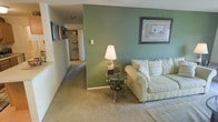 This single-floor residential space offers a welcoming and functional layout designed for comfortable everyday living. The living area serves as the heart of the home, featuring soft wall-to-wall carpeting that adds warmth underfoot. A bold sage green accent wall provides a striking focal point, complemented by framed wall art that adds a touch of character to the space. The room is well-suited for relaxed seating arrangements, as evidenced by its generous proportions. A glass-top side table paired with a classic floor lamp adds a decorative yet practical element to the living area. From the living room, a hallway extends toward additional areas of the home, lined with framed artwork that creates a gallery-like passage. The hallway leads to what appears to be a kitchen or utility area, suggesting a thoughtfully connected floor plan. Neutral-toned walls throughout the main corridor keep the space feeling bright and open. The overall ambiance of the space is cozy and lived-in, with a harmonious blend of neutral tones and earthy greens that create a calm, inviting atmosphere. The single-floor layout ensures easy accessibility and a seamless flow between living spaces.
