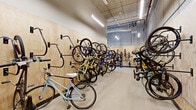 Located at 11a W Lamme St in Bozeman, Montana, this purpose-built bike storage facility offers a well-organized and generously proportioned single-level space designed to accommodate a large number of bicycles. The interior features plywood-paneled walls lined with heavy-duty black wall-mounted bike hooks on both sides, maximizing vertical storage capacity throughout the length of the room. The polished concrete flooring is durable and easy to maintain, while the exposed ceiling with linear LED lighting ensures bright, even illumination throughout the space. At one end of the facility, a dedicated repair station is equipped with a bike stand and a long wooden workbench, providing a functional area for maintenance tasks. Large steel-framed windows flood this area with natural light and offer views to the outdoors, contributing to an open and airy feel. A row of vibrant teal-colored metal lockers is situated toward the rear of the space, offering secure personal storage for cyclists. The overall design strikes a balance between industrial utility and thoughtful functionality, making this facility a practical and well-appointed amenity for bicycle commuters and enthusiasts alike.